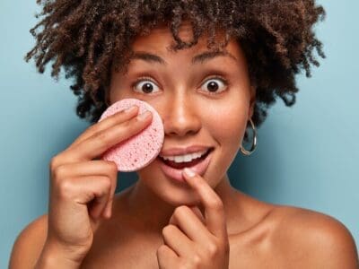 surprised cheerful afro american woman hears advice how care about skin holds cosmetic sponge cheek has widely opened eyes shocked reaction removes makeup spa relaxation concept 273609 26519